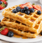 Stack of healthy almond flour waffles topped with fresh strawberries, blueberries, and pure maple syrup on a white plate for a wholesome breakfast recipe.